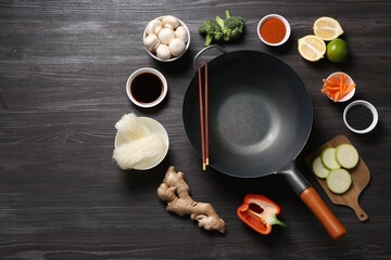 Empty iron wok and chopsticks surrounded by ingredients on dark grey wooden table, flat lay. Space for text