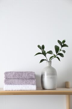 Stacked terry towels and green branches in vase on wooden shelf near white wall