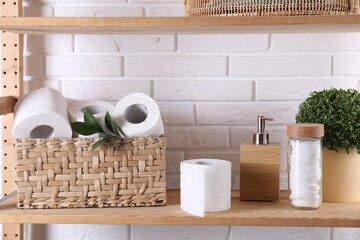Toilet paper rolls in wicker basket, floral decor, cotton pads and dispenser on wooden shelf...
