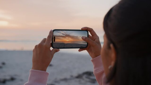 Young woman taking photo or video of sunrise at beach on mobile phone - shot in slow motion