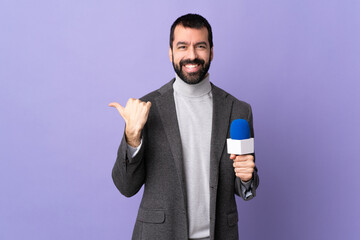 Adult reporter man with beard holding a microphone over isolated purple background pointing to the...