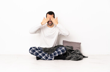 Caucasian man in pajamas sitting on the floor at indoors covering eyes by hands and smiling