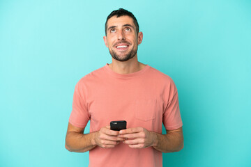 Young handsome caucasian man isolated on blue background using mobile phone and looking up
