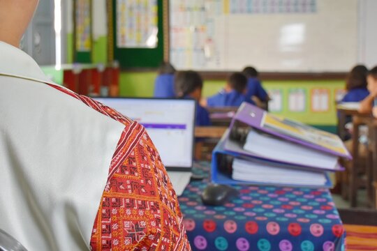 A Thai teacher is sitting and doing a lot of paperwork at a table in the classroom. While teaching at the same time.
