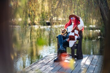 Autumn lake woman red hair dog. She sits by a pond on a wooden pier in autumn and admires nature, holding a Yorkshire terrier in her hands. The concept of tourism, weekends outside the city. - obrazy, fototapety, plakaty