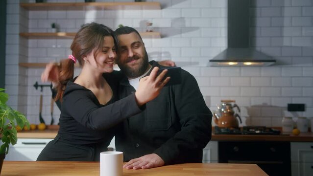 Happy young couple sitting in the kitchen taking selfie photos with smartphone smiling, having fun, having great time