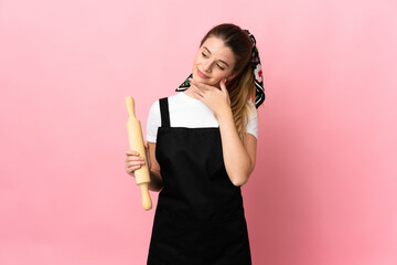 Young blonde woman holding a rolling pin isolated on pink background looking up while smiling