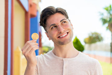 Young handsome man holding a Bitcoin at outdoors looking up while smiling