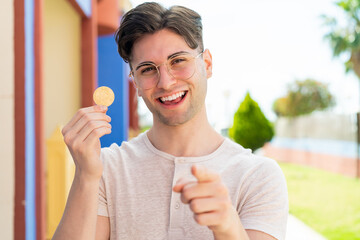 Young handsome man holding a Bitcoin at outdoors points finger at you with a confident expression