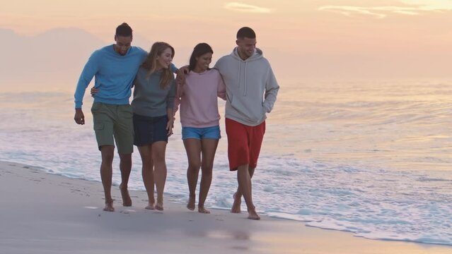 Young couple with friends wearing casual clothing hugging as they walk towards camera along beach at dawn - shot in slow motion