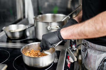 A chef in black gloves stirs ingredients in a professional kitchen. Stainless steel countertops and cookware surround them.
