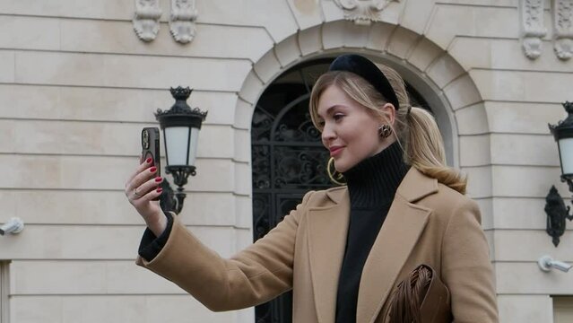 Beautiful young woman tourist takes selfie using smartphone against beautiful architecture building, traveling on historic city street. Fashionable female model in elegant clothes
