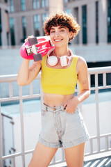 Young beautiful smiling hipster woman in trendy summer clothes. Carefree woman with curls hairstyle, posing in the street. Positive model outdoors. Cheerful and happy. Holds penny skateboard