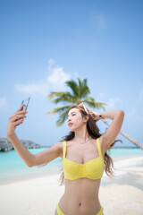Woman in yellow bikini taking selfie near the coconut palm trees on tropical beach.