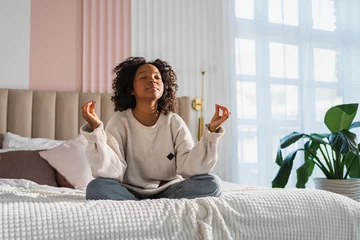 Poster Yoga mindfulness meditation. Young healthy African girl practicing yoga at home. Woman sitting in lotus pose on bed meditating smiling relaxing indoor. Girl doing breathing practice. Yoga at home © Юлия Завалишина