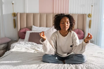 Ingelijste posters Yoga mindfulness meditation. Young healthy African girl practicing yoga at home. Woman sitting in lotus pose on bed meditating smiling relaxing indoor. Girl doing breathing practice. Yoga at home © Юлия Завалишина