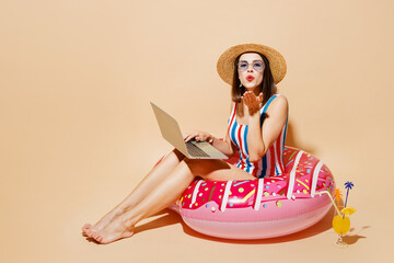 Full body young IT woman wear one-piece swimsuit straw hat near hotel pool sit on donut ring use laptop pc computer send air kiss isolated on plain beige background. Summer sea rest sun tan concept.
