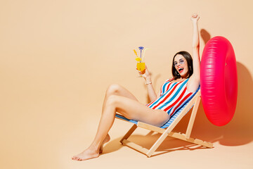 Full body young woman wear one-piece swimsuit straw hat near hotel pool sit on deckchair hold in hand orange cocktail isolated on plain light beige background Summer vacation sea rest sun tan concept
