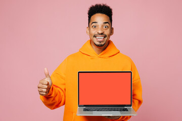 Young surprised IT man of African American ethnicity wear yellow hoody casual clothes hold use work on blank screen workspace area laptop pc computer show thumb up isolated on plain pink background.
