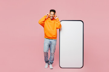 Full body young man of African American ethnicity wear yellow casual clothes big huge blank screen mobile cell phone smartphone with area listen music in headphones isolated on plain pink background.