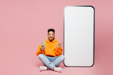Full body happy young man wearing yellow hoody casual clothes sit point finger on big huge blank screen mobile cell phone smartphone with area use mobile cell phone isolated on plain pink background.