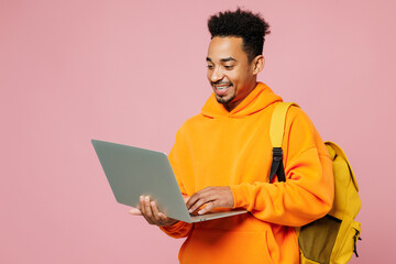 Young student IT man of African American ethnicity yellow hoody casual clothes backpack bag hold books use laptop pc computer isolated on plain pastel pink background. High school university concept.