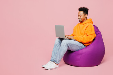 Full body young man of African American ethnicity in yellow hoody casual clothes sit in bag chair hold use work on laptop pc computer isolated on plain pastel light pink background Lifestyle concept