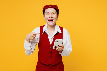 Young stewardess flight attendant woman of Asian ethnicity she in red vest shirt hat use point finger on mobile cell phone isolated on plain yellow background studio portrait Air flight trip concept