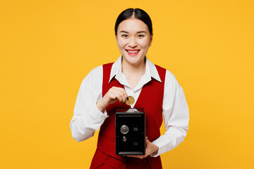 Young happy corporate lawyer employee business woman of Asian ethnicity in red vest shirt work at office hold put coin into metal bank safe for money accumulation isolated on plain yellow background