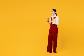 Full body side profile view young lawyer employee business woman of Asian ethnicity wear formal red vest shirt work at office hold book drink coffee walk go isolated on plain yellow background studio.