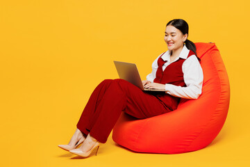 Full body young lawyer employee IT business woman of Asian ethnicity wear red vest shirt work at office sit in bag chair hold use work on laptop pc computer isolated on plain yellow background studio.