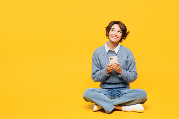 Full body young woman wears grey knitted sweater shirt casual clothes sits hold in hand use mobile cell phone look aside on area isolated on plain yellow background studio portrait. Lifestyle concept.