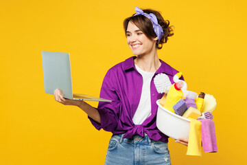Young smart IT woman wear purple shirt hold basin with detergent bottles do housework tidy up use work on laptop pc computer surfing internet isolated on plain yellow background. Housekeeping concept.