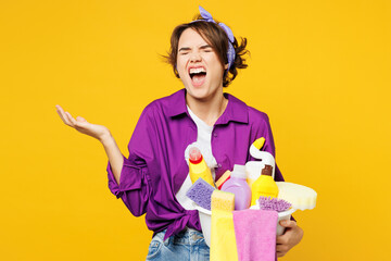 Young frustrated woman wear purple shirt hold basin with detergent bottles do housework tidy up scream spread hand close eyes isolated on plain yellow background studio portrait. Housekeeping concept.