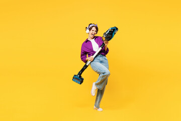 Full body young woman wear purple shirt casual clothes do housework tidy up hold vacuum cleaner listen to music in headphones pov play guitar isolated on plain yellow background. Housekeeping concept.
