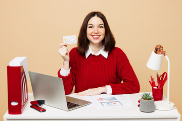 Young happy employee business woman wear red sweater shirt sit work at office desk with pc laptop hold in hand credit bank card isolated on plain pastel beige background. Achievement career concept.