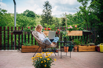 A beautiful middle-aged blonde woman in her country house is sitting on the terrace surrounded by flowers and working on a laptop. The life of downshifter in a house in nature surrounded by flowers