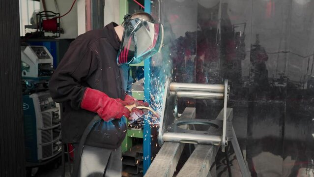 A welder in a mask welds a metal part and sparks fly in different directions.