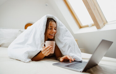 A woman is comfortably laying in bed under her blanket, with a cup of coffee and her laptop