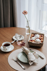 Breakfast for one person is served on a round wooden table. There is a cup with a saucer and tea, a dish with pancakes, ice cream and cherries. and a glass vase with a gerbera flower