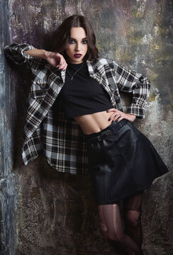 Indoor portrait of pretty grunge (rock) girl standing at wall. Gorgeous informal model, dressed in checkered shirt, topic, leather skirt and torn tights