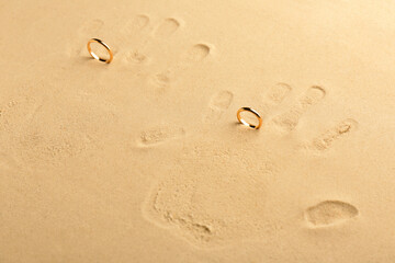 Honeymoon concept. Handprints and two golden rings on sand, above view