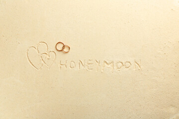 Word Honeymoon written on sand, hearts two golden rings, top view
