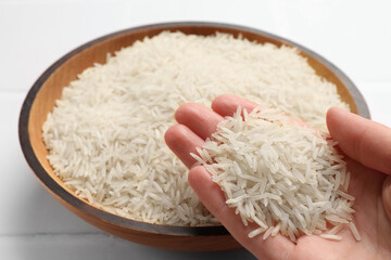 Woman holding raw basmati rice near bowl at white table, closeup