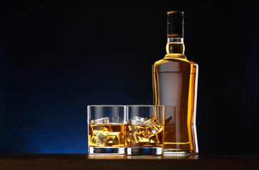 Whiskey with ice cubes in glasses and bottle on table against dark background, space for text