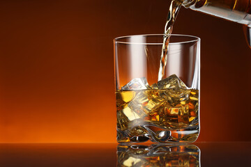 Pouring whiskey into glass with ice cubes at table against color background, space for text