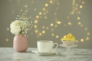 Delicious cupcake with yellow cream, tea and flowers on white marble table against blurred lights