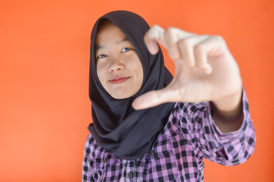 Young Asian woman in attractive purple shirt and black hijab feeling happy and romantic forming heart gesture expressing tender feelings wearing casual jumper pose on orange background. Concept of aff