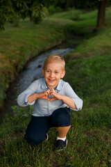 Cute little boy sitting on the grass, showing a heart sigh behind river.