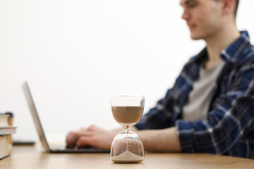 Hourglass with flowing sand on desk. Man using laptop indoors, selective focus
